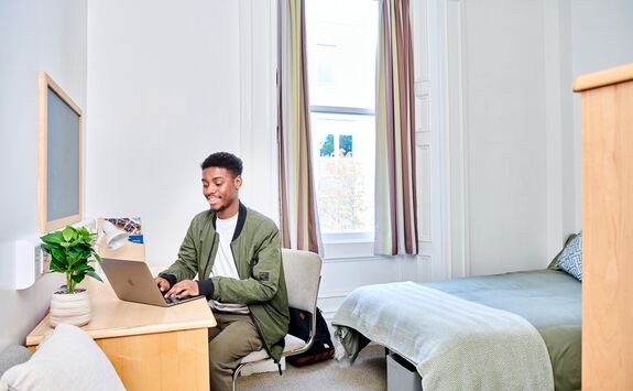 A student working on a laptop at a desk in a student accommodation room